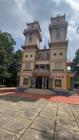 The video shows a tall, two-towered building with ornate decoration in an Asian style. The building is yellow, with blue trim, and each tower has a small roof on top. There is a balcony on the second floor, and several statues flank the entryway.  The building is surrounded by a red brick patio and a courtyard with large grey pavers.  The background includes some green trees and a cloudy sky.