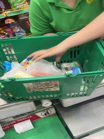 A cashier in a green polo shirt with a logo is scanning items from a green plastic shopping basket on a metal counter at a checkout. The cashier is using a handheld barcode scanner. The basket contains several plastic bags of groceries, including what appears to be produce and other packaged items. A scale is visible beneath the basket, and a keyboard and computer monitor are to the right. A receipt is visible on the counter in front of the scale. The environment appears to be a retail store.