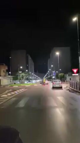 The video shows a nighttime cityscape, likely filmed from a vehicle moving along a wet street. Tall, modern buildings line both sides of the road, illuminated by streetlights. Cars and scooters are visible, moving in the same direction as the camera, as well as in the opposite direction in the distance. The sky is dark, and the overall atmosphere suggests a calm, urban night scene.