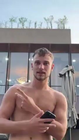 A shirtless man stands in front of a building and smiles at the camera. He's fair-skinned with short, light brown hair and a short beard. He holds a dark smartphone in his left hand. The building behind him has large windows that reflect the evening sky. The building's roof is lined with small trees or bushes. To the right of the man is a folded umbrella. The man looks happy and relaxed.