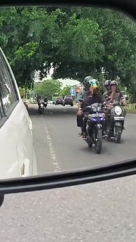 The video captures a street scene as viewed through a car's side mirror, with the white body of the viewer's car visible on the left. The setting appears to be a road in Ujong Duabelas, Indonesia, lined with abundant green trees.

On the road, several vehicles are in motion. Multiple motorcyclists are seen approaching from behind and driving past, some wearing helmets. Further down the road, a yellow dump truck is visible on the right, and other cars can be seen in the distance. The overall traffic appears moderate.

The time of day is daytime, with soft, diffused lighting suggesting an overcast sky or possibly early morning/late afternoon. No specific emotions are discernible among the visible individuals, and no explicit text is clearly legible on any signs or vehicles within the frame. The primary activity is the movement of traffic along the road.