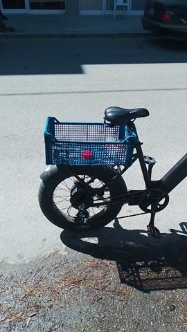 The scene shows a black e-bike with a blue crate attached to the front. The bike is parked on a paved surface, possibly a road or driveway. A car is partially visible to the left of the bike. To the right, there is a patch of gravel or dirt with some scattered leaves. The camera appears to be positioned directly above the bike, providing a bird's-eye view. The bike is in focus, while the background elements are slightly less clear. The lighting suggests it is daytime and the shot is taken outdoors. The bike's shadow is prominent on the gravel.