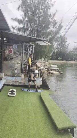 A wakeboarder in a helmet and dark shorts is positioned on a wakeboard at the edge of a green-turfed platform beside a body of water. They are bent over, seemingly preparing to start. To their left, a smaller white board with handles is on the grass. Behind the wakeboarder, a person in a yellow shirt stands on top of a metal structure, looking out towards the water. Another person is partially visible sitting on the edge of the platform on the left. In the background, trees and a cable system for wakeboarding are visible over the water. The weather appears overcast.