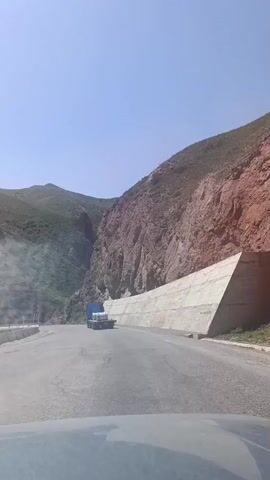 A dash cam video captures a blue truck with a flatbed trailer carrying several cylindrical objects driving on a winding mountain road under a clear sky. The road is asphalt and appears to be two lanes. On the right side of the road, a steep, reddish-brown cliff face is visible, partially reinforced with a concrete wall. The left side of the road slopes upward, covered in green vegetation, with a metal guardrail. The truck is moving away from the camera, in the direction of the road's curve.