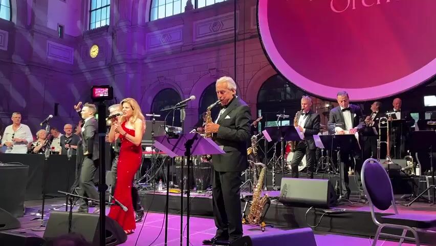 A live band performs on a stage in a large indoor venue. The band consists of a female singer in a red sequined dress, a male saxophone player in a tuxedo, and several other musicians in formal wear playing various instruments including a saxophone, trumpet, and drums. The stage is lit with purple and pink lights, and a large circular screen with a pinkish hue is visible in the background. In front of the stage, an audience is partially visible, some standing and some seated. The venue appears to be an old, grand building with high ceilings and large windows. The overall atmosphere is formal and celebratory.