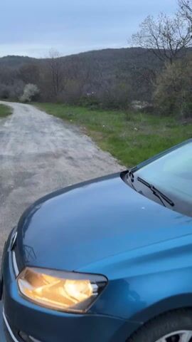 The video shows a car driving on a dirt road towards a small house with a tiled roof and a dark garage door. The house is located on the side of the road, with a grassy area around it. In the background, there is a hill covered with trees under a cloudy sky. The car is blue and the camera is positioned on the hood of the car.