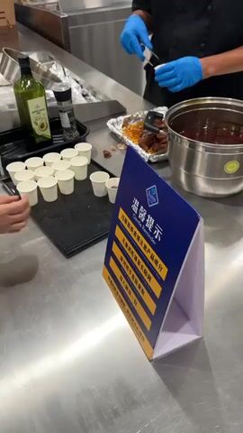 A person wearing blue gloves is serving small samples of food from a large pot into small white cups. To the left, there are several more empty white cups arranged on a black tray, a bottle of olive oil, and a pepper grinder. A sign with Chinese text and "Tasting Reminder" is in the foreground. Another person in the background appears to be preparing more food. The setting looks like a commercial kitchen or food sampling area with stainless steel surfaces.