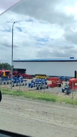 The video pans across an outdoor area filled with agricultural machinery. In the foreground, a gravel driveway is visible, with some patchy grass on the right. In the midground, various pieces of farm equipment are parked, including tractors, plows, and what appear to be harvesters or rakes. Some of the machinery is red, green, and yellow. A long, white building with a blue roof stands in the background. Two tall poles with lights or cameras are positioned on either side of the frame. The sky is cloudy.