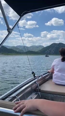 This video captures a scenic boat ride on a sunny day, likely in the Prevlaka region of Montenegro. The camera is positioned from within a boat, looking out over the water. In the foreground, we see the edge of the boat with metal railings and seating, as well as parts of two people's hands and a woman's shoulder and back wearing a light pink shirt.

The main subject of the video is the expansive body of water, which appears to be a lake or a calm bay, reflecting the bright blue sky dotted with fluffy white clouds. In the middle ground, another boat is visible, moving away from the camera. This boat has a canopy and appears to be carrying several passengers. Further in the distance, a range of rolling, verdant hills forms a picturesque backdrop, suggesting a lush, natural environment.

The overall mood is peaceful and leisurely, indicative of a pleasant outing on the water. The clear skies and calm waters suggest fair weather, making it an ideal day for boating. The presence of other boats and the natural landscape point towards a popular tourist or recreational area. Visible on the distant boat is text that reads "RBBINA JELENA". The time of day appears to be midday, given the bright sunlight.