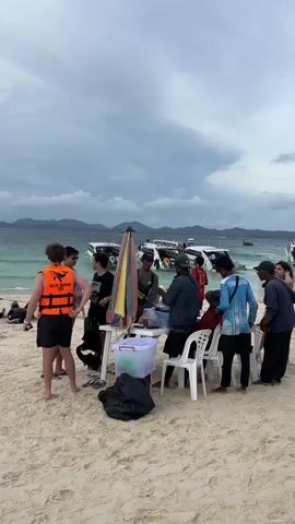 The video shows a group of people on a sandy beach, with the ocean and boats in the background. A man in an orange life vest stands on the left, facing away from the camera. To his right, several people are gathered around a table set up with a colorful umbrella. Some are standing, while others are seated on white chairs. Objects on the table include a clear plastic bin with a red lid, and what appears to be black bags on the ground. Several individuals are interacting with each other, with some appearing to be engaged in conversation. The water is choppy, and the sky is overcast with clouds. Several speedboats are docked or anchored in the water.