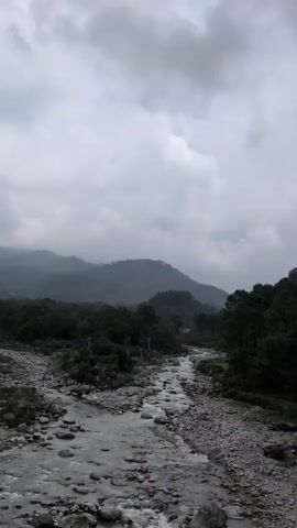 A wide shot shows a mountain landscape under a cloudy sky. A rocky river flows through the center of the frame, with dense green trees on either side. In the background, rolling hills are shrouded in mist, with the peaks disappearing into the clouds. The overall atmosphere is calm and serene.