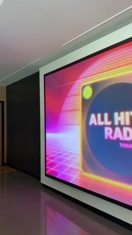 The video shows a large screen displaying a retro-themed graphic with neon grids. The graphic transitions between two images. The first image features the band Imagine Dragons with the text "ALL HITS RADIO SPECIAL GUEST: IMAGINE DRAGONS". The second image shows a woman with the text "ALL HITS RADIO SPECIAL GUEST: MAROON 5". The screen is mounted on a wall in a room with a white frame around it. The room appears to be a modern space with dark walls and a tiled floor. The lighting suggests an indoor setting.