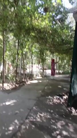 The video shows an outdoor scene with a path covered by an overhead structure of vines and leaves. A person in a red and white patterned dress is walking away from the camera under the canopy. Another person in a white dress is further down the path, also walking away from the camera. The path is made of concrete and is partly shaded by the foliage. Trees and greenery are visible on the sides of the path. The overall environment appears to be a garden or park area.
