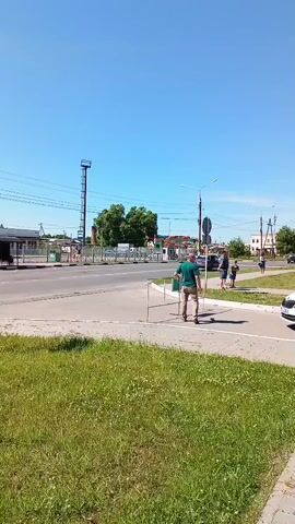 The video shows an outdoor scene on a clear, sunny day. A man in a green shirt and khaki pants walks left across a grassy foreground on a paved sidewalk, carrying several white, thin objects in his right hand. A circular road sign on a pole is positioned to the left of the man. In the background, a two-lane road features several vehicles moving from left to right. A dark gray SUV drives past, followed by another dark gray SUV. Subsequently, a white police car with blue and white stripes drives past, followed by a yellow minibus. On the far side of the road, a group of people are gathered on a sidewalk. Buildings and trees are visible further in the background, along with power lines and a tall metal tower-like structure on the left side of the frame.