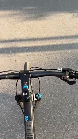 The video offers a first-person perspective from someone riding a bicycle. The camera is mounted on the bike, looking down at the handlebars and front wheel, with the road ahead visible.

The primary subject is a black mountain bike, clearly identified by the visible text "ROCKRIDER 520" on its frame. The handlebars, stem (with a blue accent), front brake levers, gear shifters, and suspension fork are all in view. Small technical markings, such as "6 NM," can be seen on the stem. The front wheel and tire are also visible, moving forward.

The bike is moving along a paved asphalt road, which appears to be a street or path. Long, distinct shadows are cast across the road, suggesting either early morning or late afternoon lighting, indicating sunny weather. The light has a warm hue. The activity depicted is simply riding the bicycle.

The video is set in Brenitsa, Bulgaria, although no specific landmarks confirming this are visible within the frame. The overall impression is a casual bike ride on a clear day.