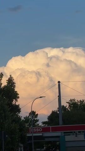 The video is a vertical, panning shot of a large, fluffy cumulus cloud illuminated by sunlight, appearing yellow and orange. Below the cloud, silhouetted trees and what appears to be the roof of a building are visible. Overhead wires and a lamppost are also present in the foreground. The sky above is a clear blue. The video is brief and focuses on the visual spectacle of the cloud.