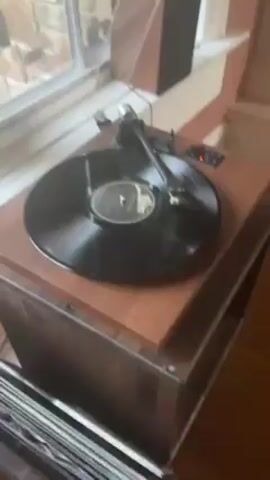 The video shows a record player on a wooden stand, playing a vinyl record. A speaker sits on the window sill behind the turntable.  A stack of vinyl records rests on the floor next to the stand. The walls are painted in a light peach color, and there are hardwood floors. Natural light filters through the window.