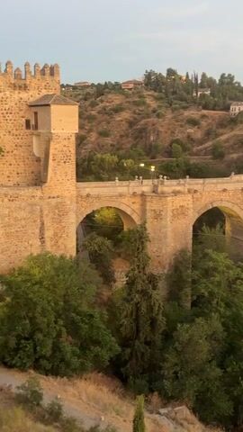 This video shows a stone bridge with multiple arches spanning a body of water. The bridge appears to be old and is made of weathered stone. There is a tower structure at the far end of the bridge. The surrounding area is hilly and covered with vegetation, including trees and bushes. Buildings are visible on the hillsides, and streetlights are illuminated, suggesting it is either dusk or early evening. The water below reflects the lights from the bridge and the buildings. The video pans slowly to the right, revealing more of the bridge and the town.