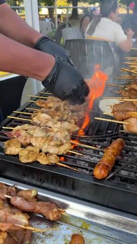 A person wearing black gloves is shown grilling skewers of meat over an open flame. The grill is a black metal grate with charcoal underneath. There are many skewers of meat on the grill, some are chicken and some appear to be sausage. The person in the black gloves is holding a small green bottle and appears to be pouring a liquid onto the grill, which is causing smoke and flames to erupt. In the foreground, there is a metal tray filled with more skewers of uncooked meat. In the background, there are people sitting at tables outdoors, and trees and foliage can be seen. The overall setting appears to be an outdoor food stall or event.