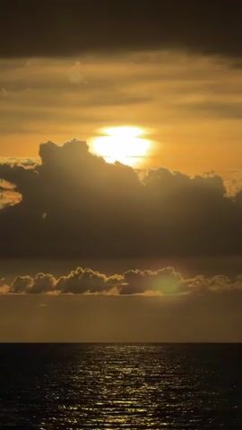 This media file captures a serene sunset over the ocean near Baan Bang Rak, Thailand. The scene is dominated by a dramatic sky filled with golden and dark clouds, with the sun peeking through, casting a brilliant, shimmering reflection on the water's surface. The ocean appears calm, with gentle ripples catching the light.

On the left side of the frame, a wooden pier or jetty juts out into the water, adding a structural element to the natural landscape. No people are visible in the media, contributing to the peaceful and solitary atmosphere.

The time of day is clearly sunset, as indicated by the warm, low-angled sunlight and the rich, colorful hues in the sky. The weather appears to be partly cloudy, with the clouds playing a significant role in shaping the dramatic lighting. The location cues, like the style of the pier and the tropical feel of the water and sky, strongly suggest a coastal setting in Southeast Asia, consistent with Baan Bang Rak, Thailand.

The overall mood conveyed is one of tranquility, beauty, and natural grandeur. The interplay of light and shadow, the vastness of the ocean, and the vivid colors of the sunset create a visually stunning and emotionally resonant image. There is no visible text within the media file.