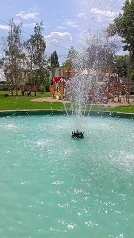 A fountain sprays water in a turquoise pool. In the background, a playground with colorful structures and slides is visible, surrounded by green grass and trees. The sky is blue with scattered clouds. The scene appears to be a park or recreational area on a sunny day.