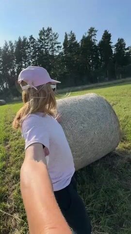 A person's arm extends into the foreground, holding a camera, facing a young woman wearing a pink baseball cap and a pink t-shirt. She is leaning against a large, round bale of hay in a grassy field under a clear blue sky. The woman turns her head to look at the camera and smiles. In the background, there are trees and another hay bale. The perspective suggests the person holding the camera is taking a selfie-style video with the woman.