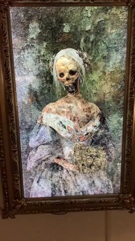 The video shows a framed portrait with a zombie bride. The portrait is lit from behind, and the zombie bride is depicted with decaying flesh, exposed bones, and empty eye sockets. She is wearing a tattered white wedding dress and holding a bouquet of flowers. The background of the portrait is a mottled green and gray, resembling a decaying forest. The frame is ornate and gold-colored. The video pans slightly, showing the portrait from different angles. The overall impression is that of a Halloween decoration or a piece of macabre art.