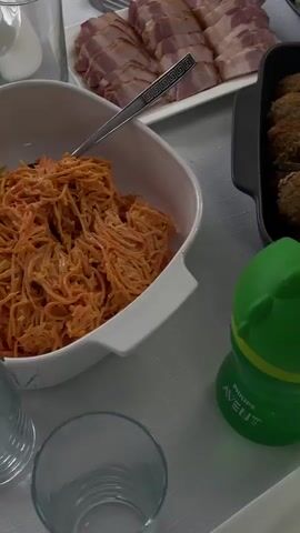 The video pans across a table laden with food, set for a meal. In the foreground, a green baby bottle with a yellow top is placed on the table. To the left, a white dish contains shredded orange food, possibly carrots. Behind it, a silver serving dish holds sliced cured meat, likely bacon. Further back, a black dish is filled with numerous brown meatballs. A larger silver catering dish in the background is overflowing with what appears to be stuffed cabbage rolls or similar dishes. A bowl of scrambled eggs is visible on the upper right. Several glasses and white plates are arranged on the table. The setting suggests a meal is about to be served or has just begun.