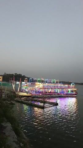 A video shows a body of water with several boats. The sky is overcast. In the foreground, a large boat is docked, decorated with colorful lights that reflect on the water. To the right and in the background, other boats with lights can be seen on the water, and a distant shoreline with hills is visible.