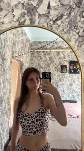 A young woman with long brown hair is standing in front of a mirror and taking a selfie. She is wearing a leopard-print crop top and matching shorts. She is also wearing pink sunglasses. The room she is in appears to be a bedroom, with a bed and a dresser visible in the background. The walls are covered in a patterned wallpaper.