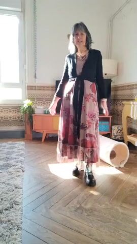 A medium shot shows a middle-aged woman standing on a wooden floor, modeling. The woman has medium length, gray hair, and fair skin. She is wearing a long floral dress, a black cardigan, and black boots. The dress has a floral pattern with reds, pinks, and tans. The cardigan is tied in the front with a black sash. The woman turns and poses, displaying the outfit from various angles. Behind her is a television on top of a retro-style cabinet, a window with natural light coming through, and a shag rug on the left side of the frame. A cat tunnel is on the floor to the right of the woman. The walls are decorated with patterned wallpaper.