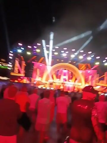 The video shows a crowd of people gathered in front of a stage at night. The stage is brightly lit with various colors, including pink, orange, and blue. There are spotlights shining into the crowd. The stage has a large arched structure in the center with heart-shaped designs. The crowd is mostly composed of people facing the stage, some wearing light-colored shirts and shorts. One person in the foreground is wearing a black sequined vest and a baseball cap. Buildings and lights can be seen in the background, suggesting an urban environment. The overall atmosphere is lively and celebratory, indicating a concert or event taking place.