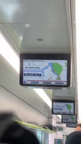 The video shows a close-up view of a digital display screen mounted on the ceiling of a train car. The screen displays a map of a body of water and surrounding land, with a route marked by a red line. The train's current location is indicated by a red circle on the map. Below the map, the screen shows the train operator's logo, "TRENORD", the current station, "Mandello Del L.", and the scheduled arrival time, "14:46". The train is in motion, as evidenced by the slightly blurred background and the movement of the train car. In the background, several other identical display screens are visible, each showing the same information. The environment appears to be clean and modern, typical of a contemporary train interior. No people are clearly visible in the foreground, but the presence of the screens and the train setting suggest passengers are on board.