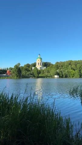A vertical video shows a serene lake with tall grass in the foreground, gently rippling water reflecting the clear blue sky. Across the lake, a white building with a green dome, possibly a church or a historic structure, sits atop a hill surrounded by lush green trees. A small red building is partially visible to the left of the main structure. The scene appears to be set in a tranquil natural environment during daylight hours. No people are visible in the video.