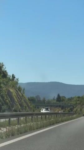 The video shows a view from a car driving on a highway. The road curves to the right. On the left side of the road, there is a steep embankment covered in grass and trees. A metal guardrail runs along the side of the road. In the distance, there are hills covered in trees. A bridge crosses over the highway. Several cars are visible on the highway in front of and in the distance. The sky is clear and blue.
