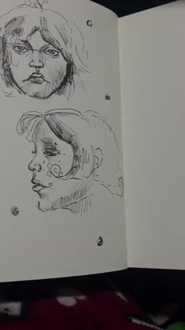 The video shows a close-up of a sketchbook page with several ink drawings of faces. The drawings are in a sketchy, somewhat loose style. The page is white and appears to be from a spiral-bound sketchbook. Small, circular doodled elements are scattered around the faces. The lighting is dim, suggesting the photo was taken in low light conditions. The bottom of the image shows a hint of a dark, textured surface, possibly fabric. The drawings themselves are the primary focus, showcasing different facial expressions and features.