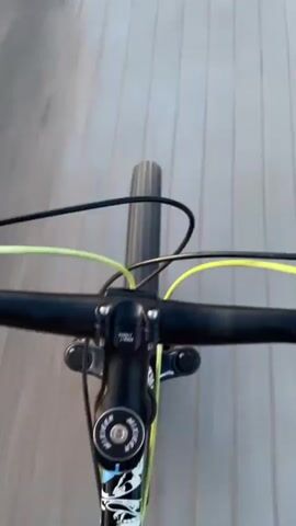 The video shows a first-person perspective of someone riding a bicycle on a wooden path. The path is wet, with puddles visible. The camera is pointed downwards, showing the front wheel of the bicycle. The rider then transitions to riding on a gravel road. The surroundings are fields of grass and trees. Power lines and poles are visible in the distance. The sky is a clear blue with a hint of orange and yellow on the horizon, suggesting it is either sunrise or sunset. The motion is consistent with cycling.