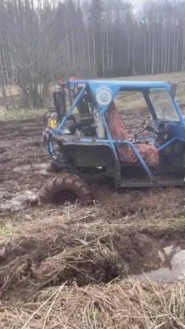 The video shows two off-road vehicles stuck in muddy conditions. The first vehicle, a blue utility terrain vehicle (UTV), is shown partially submerged in a mud pit. The video then pans to a wider view showing more muddy terrain, other vehicles, and people wearing white protective suits. The video then cuts to a white and black Niva, also stuck in deep mud, with mud caked on its wheels and body. A person is standing near the open driver's side door.