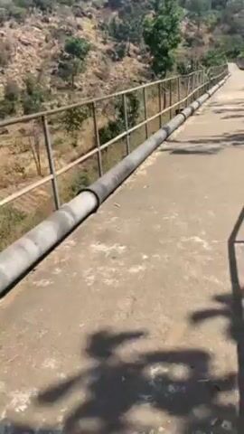 The video shows a concrete walkway with metal railings and a pipe running along it, leading toward a mountain. The walkway is cracked and worn. The mountain is covered in dry grass, rocks, and sparse trees. The sky is blue with scattered clouds. The overall impression is one of a rural or semi-arid landscape.