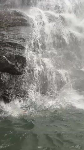 A waterfall cascading over dark rocks fills most of the frame. The water is white and foamy, creating a misty atmosphere. At the base of the waterfall, the water appears to form a small pool. Briefly, two people's heads appear in the water, then disappear. The camera remains focused on the waterfall and pool.