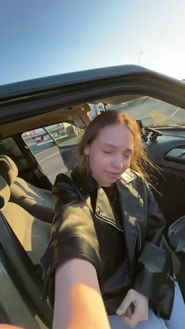 A woman in a black leather jacket sits in a parked car on a sunny day. A man exits the car and begins dancing. He is wearing a gray t-shirt and black pants. The car is parked on a curb next to a field. There is a truck parked in the background.