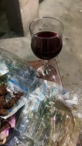 A glass of red wine sits on a wooden surface next to a pile of fried meat and onions, which is wrapped in newspaper. The scene appears to be outdoors, with a concrete surface visible in the background. The lighting suggests it is evening or nighttime.
