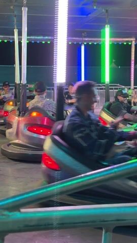 The video shows several people riding bumper cars at a fair or amusement park. The cars are various colors, including red, silver, and blue, and have lights on them. The riders are wearing casual clothing and appear to be enjoying themselves. The ride is surrounded by a metal railing, and the area is lit with colorful lights. The camera moves slightly, capturing the bumper cars bumping into each other.