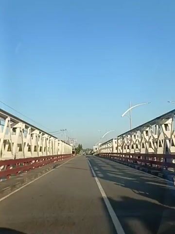 The video shows a view from inside a vehicle traveling on a road that crosses a bridge. The bridge has white trusses with red railings. The road has a white line marking the center and the right edge. The sky is clear and blue. There are power lines and streetlights visible along the sides of the road. In the distance, there are buildings and trees. The vehicle appears to be moving forward, as indicated by the perspective of the road and the slight motion blur. There are no other vehicles or people clearly visible in the immediate vicinity. The overall scene is one of a typical commute or travel on a bridge in daylight.