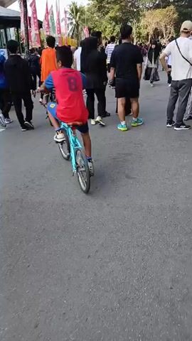 The video shows an outdoor scene with a group of people, some walking and others on bicycles. A young boy wearing a red shirt with the number "8" on the back is riding a small, light blue bicycle in the foreground. He is facing away from the camera. To his left, a person in a blue shirt and dark pants is walking. To his right, a person wearing a black hijab and a black outfit is standing, and behind them, others are walking or standing. In the background, more people are visible, some walking and others sitting. There are also some colorful banners or flags on poles. The ground is paved asphalt. The lighting suggests it is daytime, possibly late afternoon due to the angle of the light and shadows. The overall atmosphere appears to be casual, possibly a community event or a gathering. The camera movement is slightly shaky, indicating it was likely handheld.