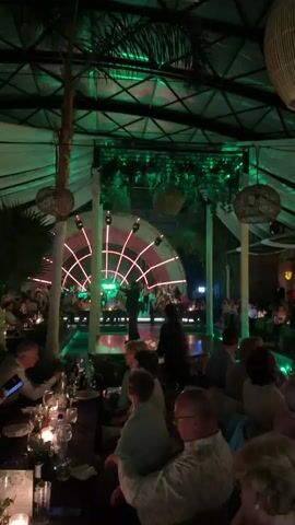 The scene takes place at an indoor event space with a performance stage. There are people sitting at tables, and a performance is ongoing on the stage. The space is adorned with lighting, including stage lights and decorative lanterns. A palm tree is also visible indoors. The event appears to be either a dinner theater or a special performance hosted in a restaurant or event hall.
