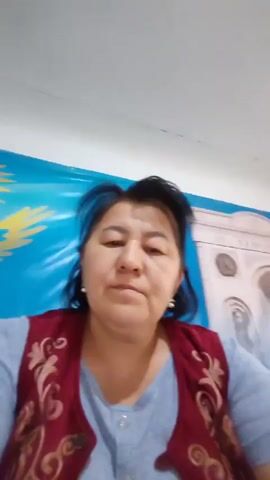 The video shows a medium shot of a woman sitting at what appears to be a desk, likely at work. She is facing the camera directly and speaking. The woman has dark hair and is wearing a light blue shirt and a dark red vest with ornate gold embroidery. She is also wearing pearl earrings. Behind her, there is a blue wall with a yellow sun-like design on the left and a partially visible structure that resembles a building facade on the right. The woman gestures with her hands as she speaks, and her facial expressions indicate that she is engaged in a conversation or giving a report. The audio suggests she is discussing being at work after a period of absence due to illness.