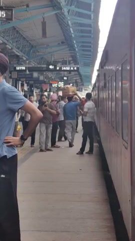 A busy train station platform. People are waiting for trains, some carrying bags and packages. One man is carrying a large box on his head. The train is visible on the right side of the frame. The time on the station display reads 14:52.