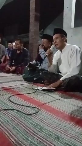 The video shows a group of men sitting on a woven mat indoors, likely at night as the lighting appears to be artificial. In the foreground, a man in a white long-sleeved shirt and a black peci (cap) is holding a microphone and singing or speaking into it. He has a book open in front of him on the mat. To his left, another man in a blue shirt and a black peci is also holding a microphone, and appears to be laughing. Further left, another man in a patterned shirt is seated. The background is dark, with some pillars and a wall visible. The overall atmosphere seems to be a gathering or performance.