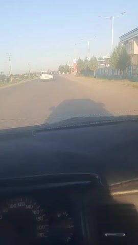 The video shows a first-person perspective from inside a car driving on a road. The car's dashboard is visible, including a speedometer. The road is asphalt and appears to be in a rural or suburban area, with trees and buildings lining the sides. There are other cars on the road, including a white car ahead and a silver car further in the distance. A gas station with a tall sign is visible on the right side of the road. The sky is clear and blue, suggesting it is daytime. The lighting indicates it might be late afternoon or early morning.